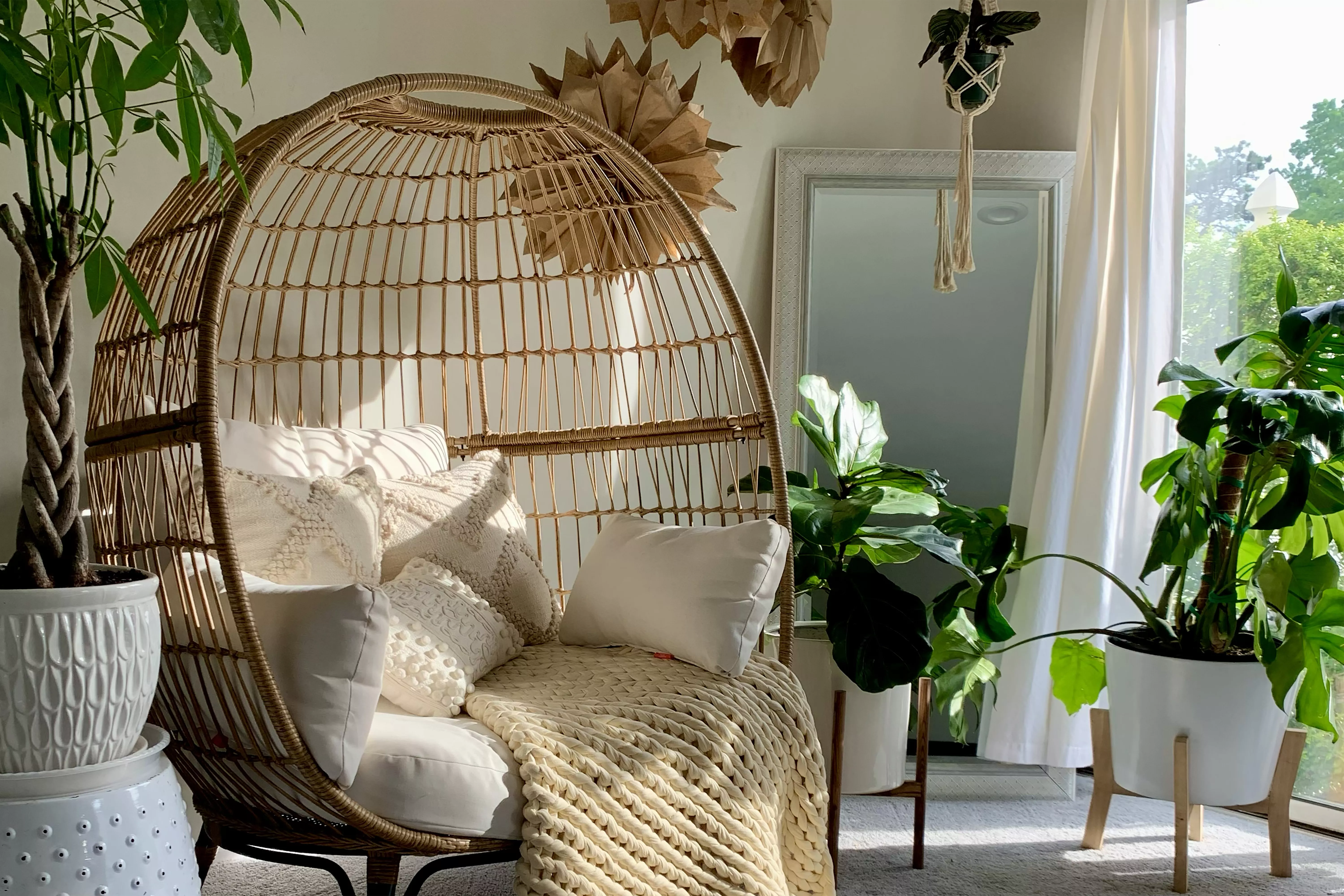 A cozy corner with a wicker egg chair, cushions, a blanket, and surrounded by potted plants